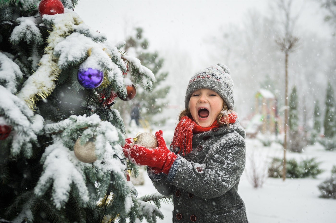 Child in winter clothes decorating a snowy Christmas tree outside